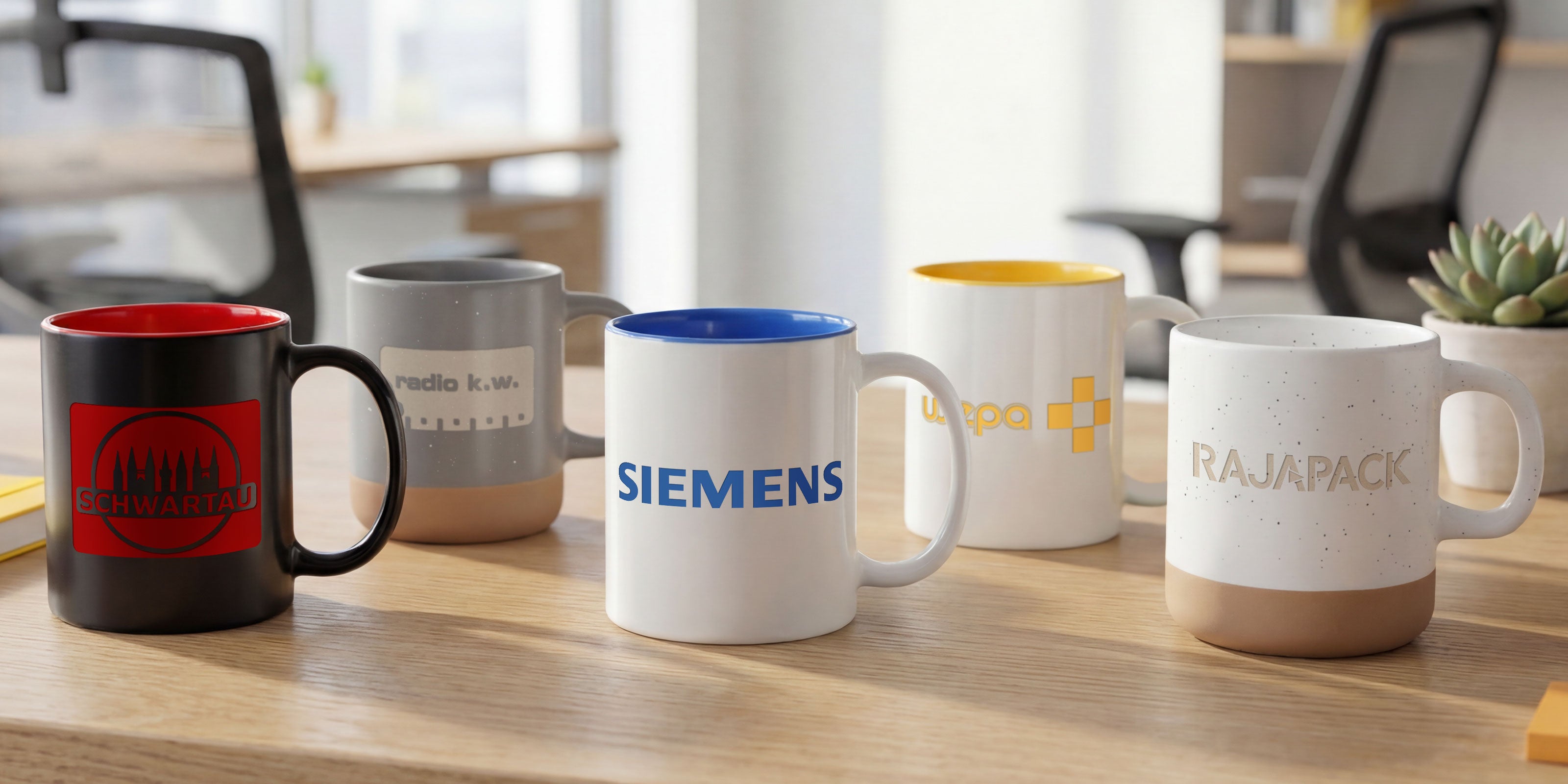 5 lasergravierte Tassen stehen auf einem Tisch in einem lichtdurchfluteten Büro. Die Tassen sind offensichtlich mit namhaften Firmenlogos gelasert worden wie Schwartau, radio k.w. Siemens, Wepa und Rajapack.